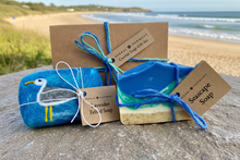 Load image into Gallery viewer, Two pieces of blue coloured soap, one is felted with a seagull, one depicts a beach and ocean waves. They are next to decorative packaging on a sandy surface near the ocean.