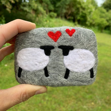 Load image into Gallery viewer, Hand holding a felted soap which is felted with light grey sheeps wool, with two sheep facing each other and two red hearts above them, against a green outdoor background. Ideal wool gift for husband or wife for 7th wedding anniversary.
