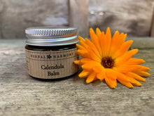 Load image into Gallery viewer, A glass jar with an aluminium lid labeled 'Calendula Balm' by Herbal Harmony, accompanied by an orange calendula flower, on a wooden surface. Handmade and natural skincare made with Calendula infused oil, beewax, lavender & tea tree essential oils.