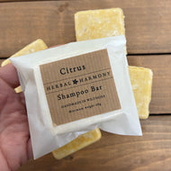 Handmade square shaped citrus shampoo bar by 'Herbal Harmony' on a wooden background. The bars are yellow coloured from yellow clay and packaged in an Eco friendly glassine paper bag. 
Natural, vegan and plastic free haircare. 
Citrus shampoo bar is scented with May Chang, orange and lime essential oils and the active ingredients include Panthenol, argan oil, jojoba oil, cocoa butter and coconut oil. 